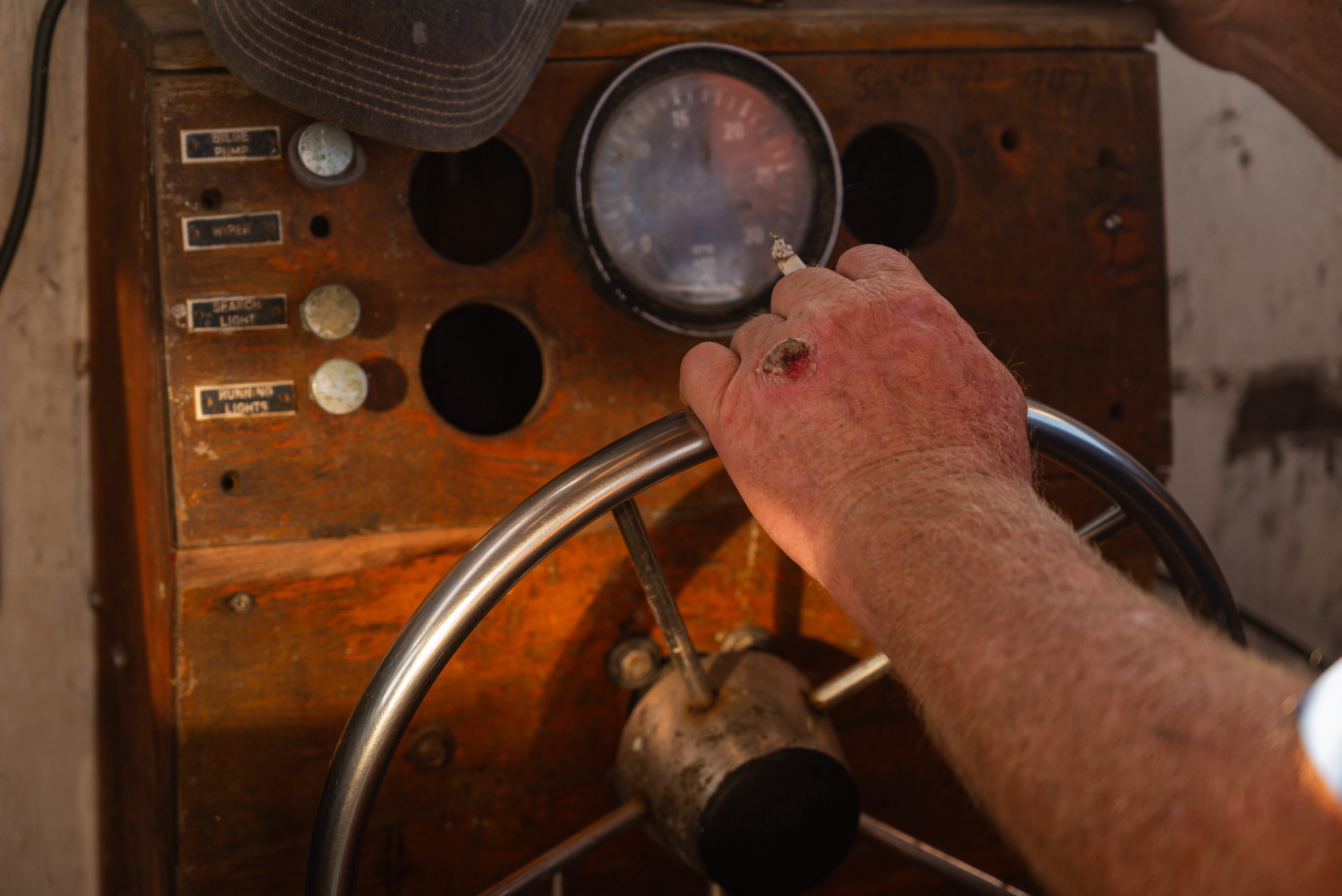 A waterman's hand rests on the helm wheel of his workboat, cigarette between his fingers.