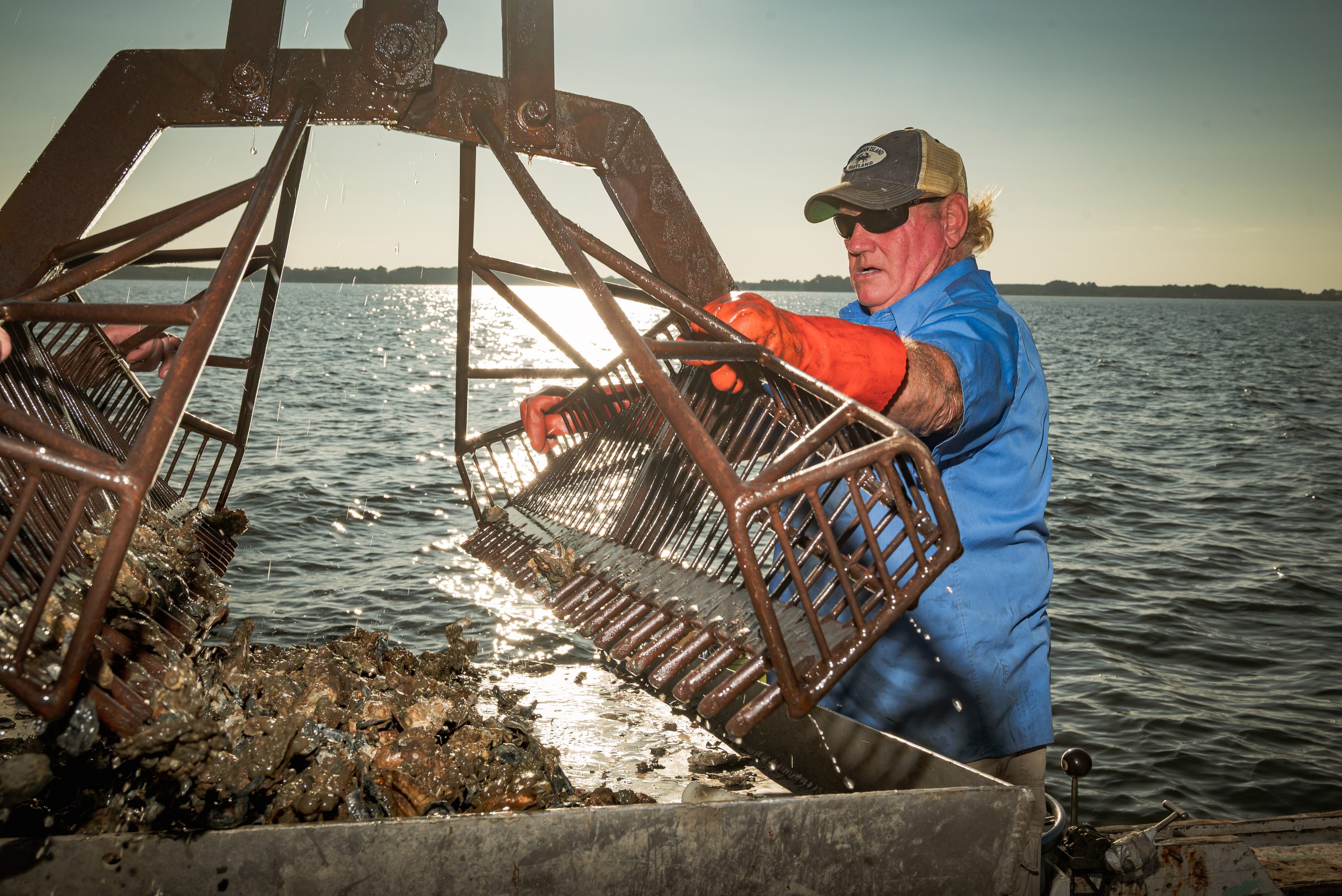 A waterman stands aboard his workboat on calm bay water in early light.