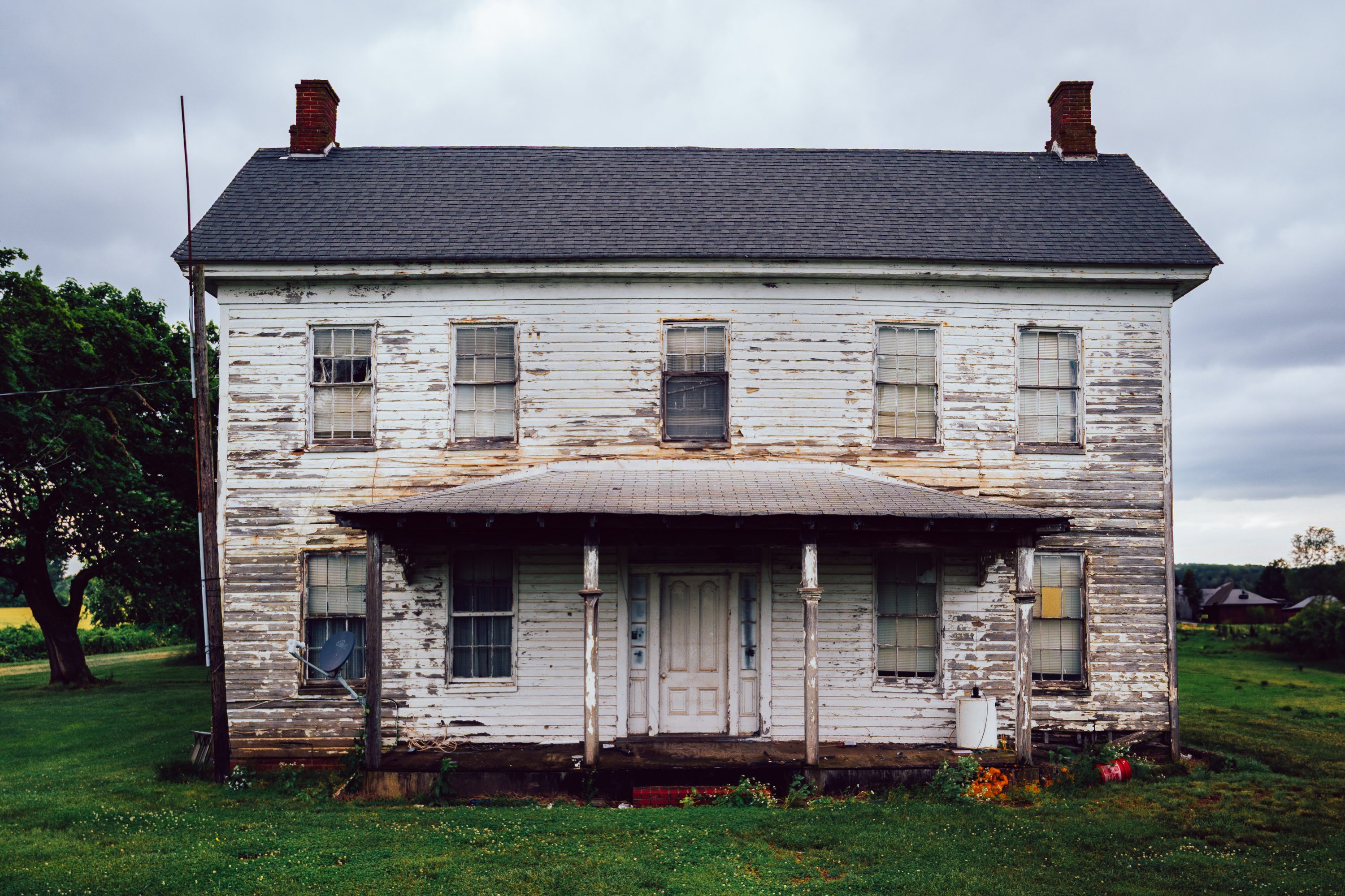 A white two-story farmhouse stands weathered against a grey sky.