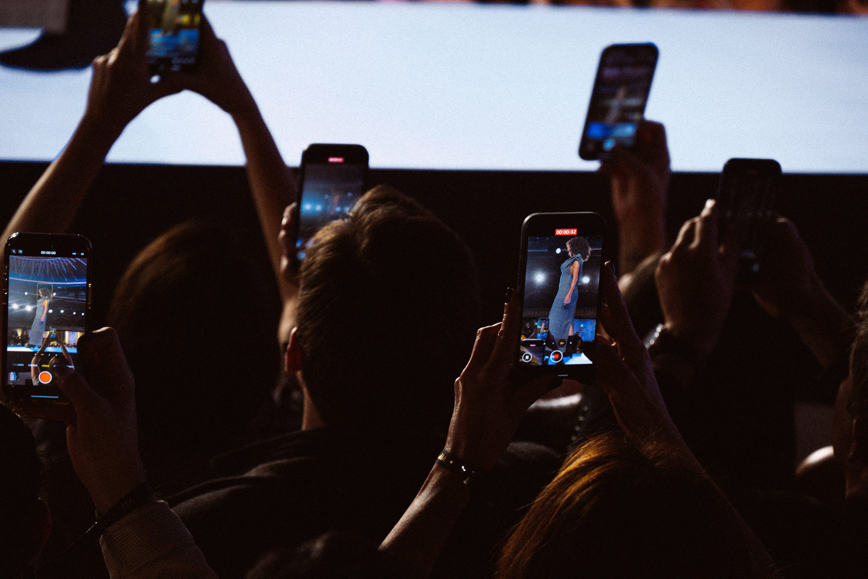 Audience members raise their phones toward the runway in deep red light.