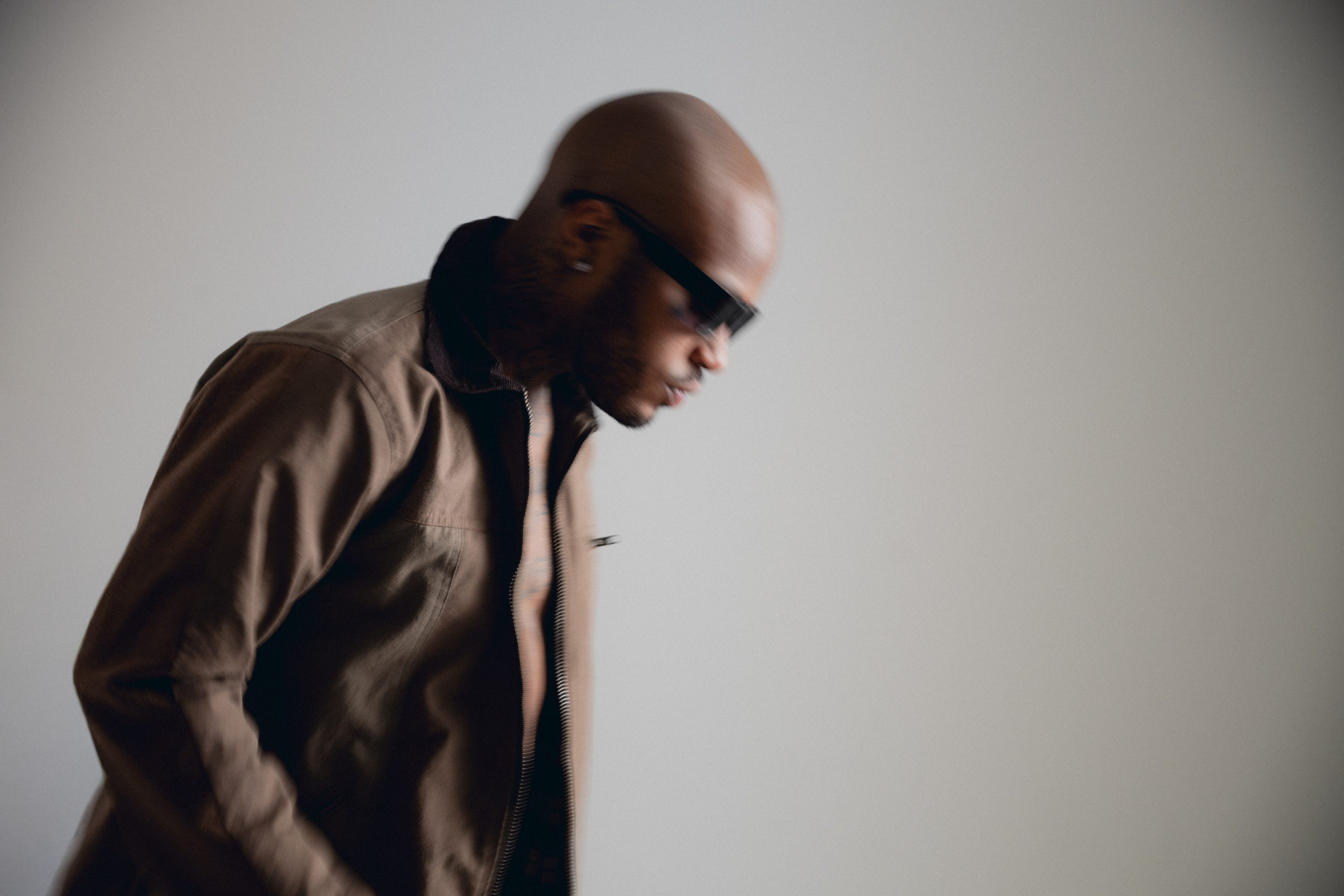 A man in sunglasses and a brown coat stands against a grey studio wall.