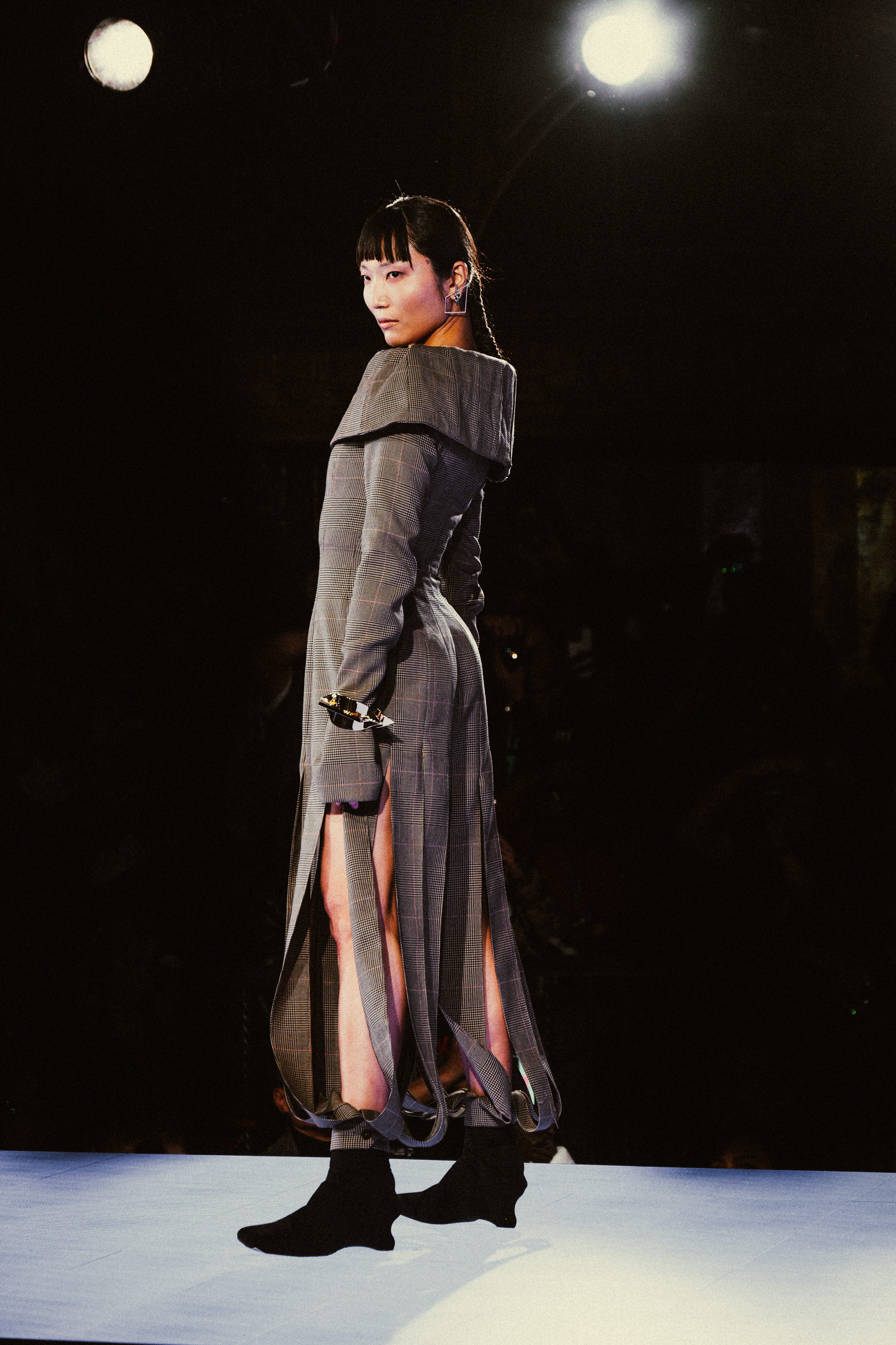 A model in a sheer layered dress walks the runway under a single spotlight.