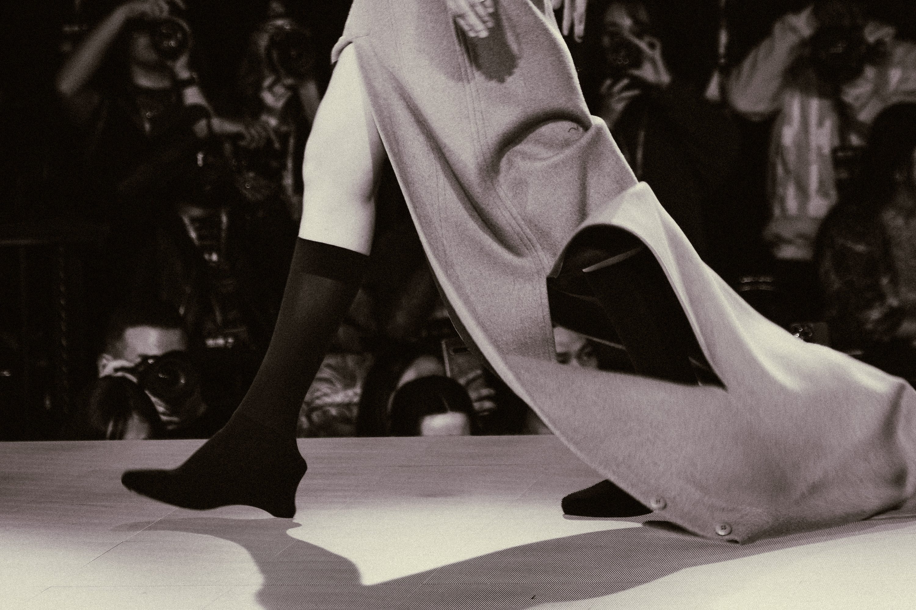 A garment sweeps behind a model mid-stride on the runway, in sepia tones.