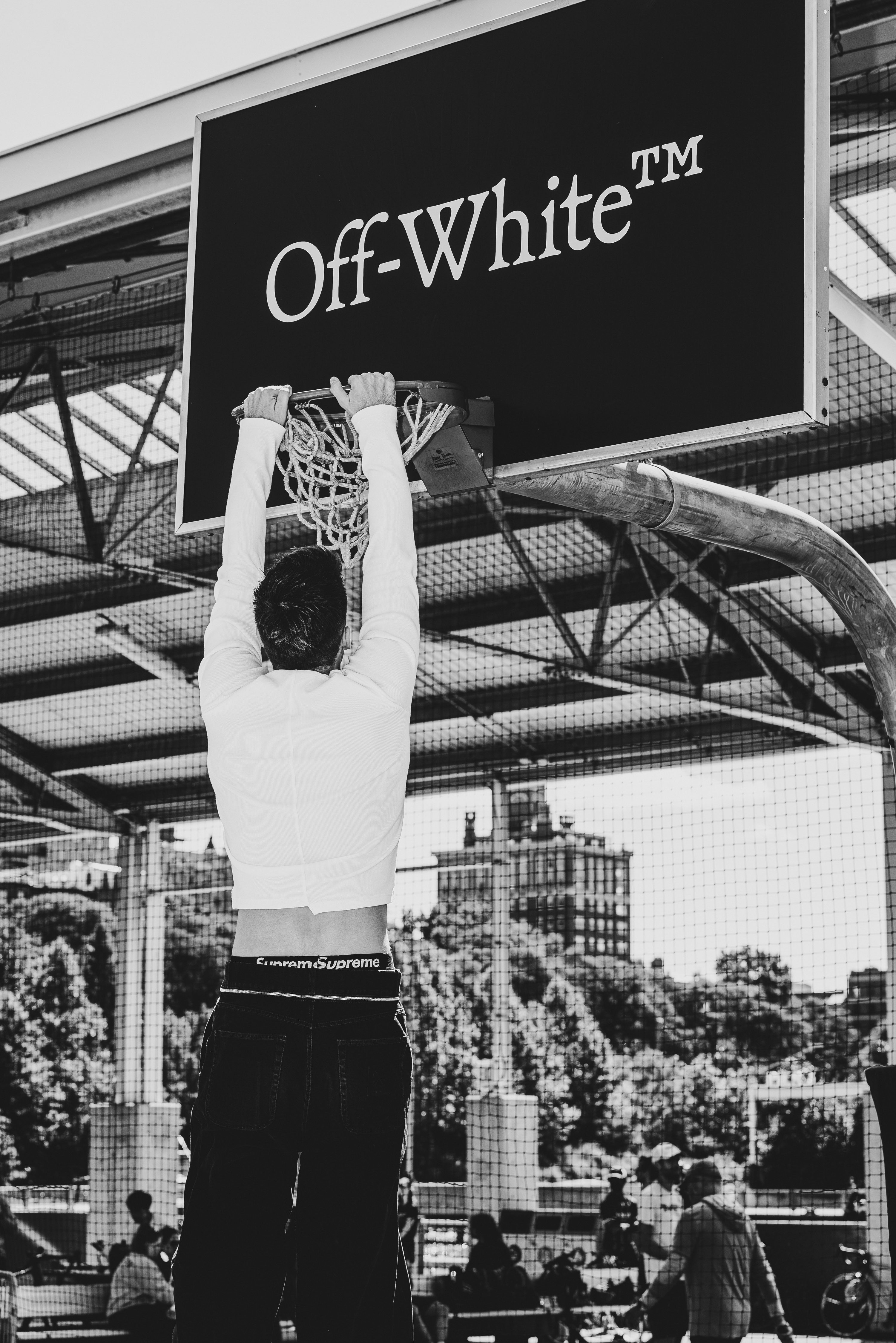 A figure hangs from an Off-White backboard sign above an outdoor court, in black and white.