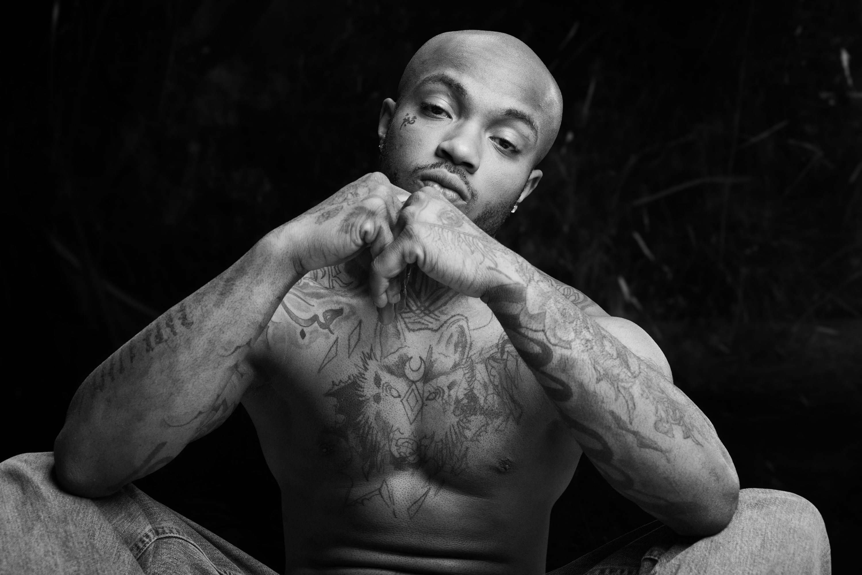 A tattooed man sits shirtless with hands clasped, photographed in black and white.