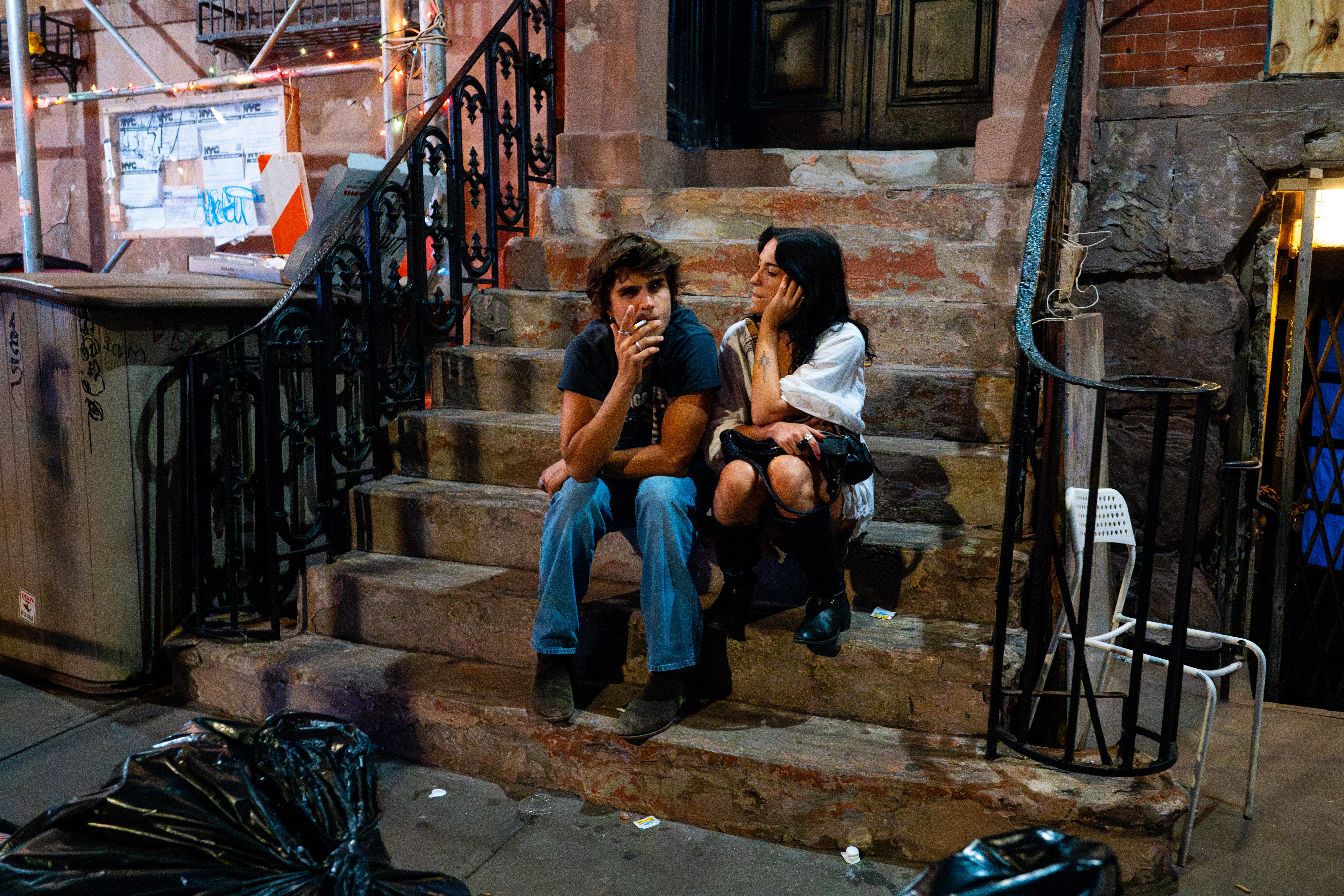 A young couple shares a quiet cigarette on the steps of a New York stoop at night.